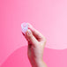 Käsi pitelee Kuukautiskuppia vaaleanpunaisella taustalla.