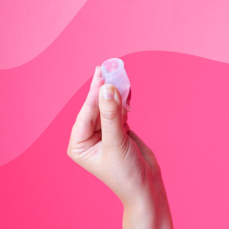 Käsi pitelee Kuukautiskuppia, pientä, läpikuultavaa silikonikuppia.