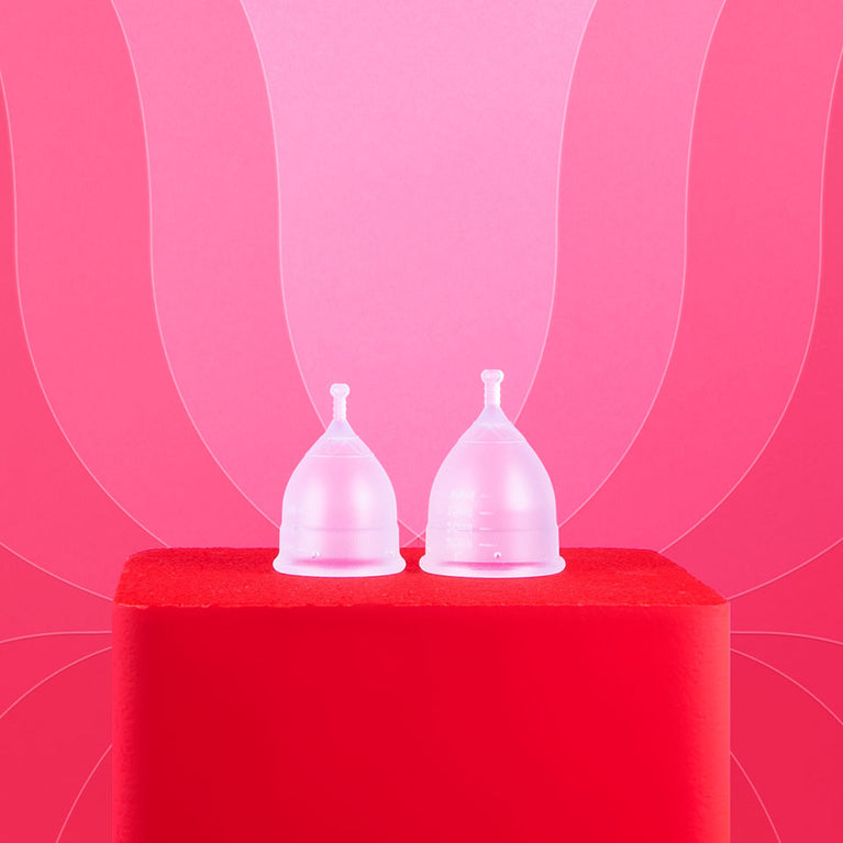 Kaksi läpikuultavaa Kuukautiskuppia punaisella alustalla.